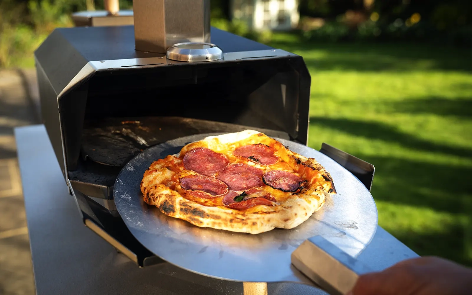 Close-up of a cooked pizza being taken out of a pizza oven on a pizza peel in a garden