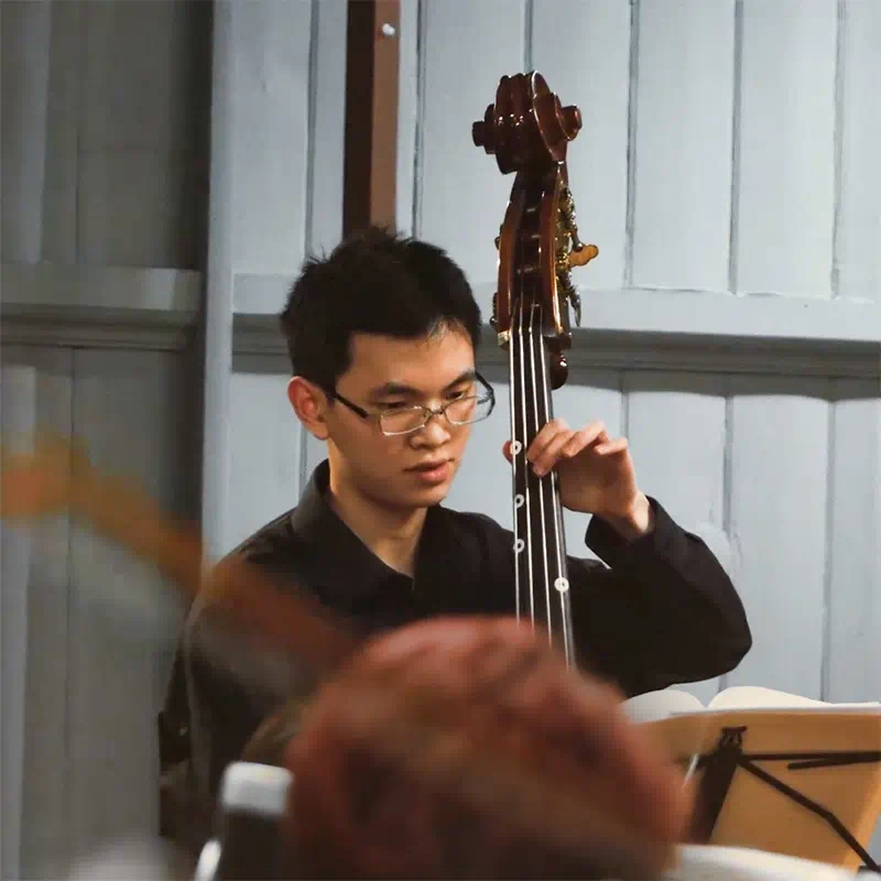 A still from the Queen Mary Music Society promotional video showing a young man playing a double bass in the orchestra