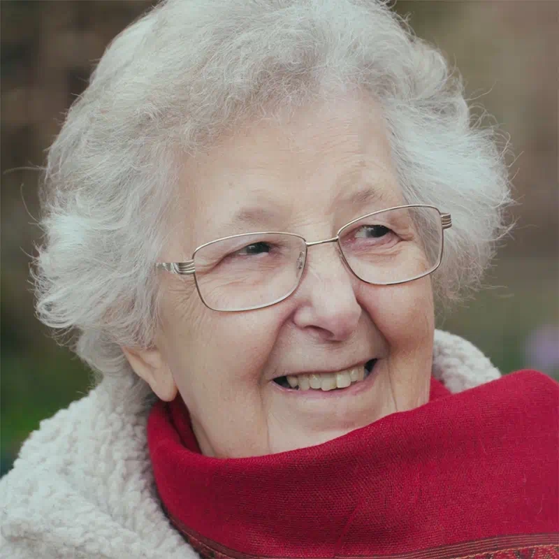 A still from the St John’s, Winchester promotional video showing a close up of an elderly lady, wearing glasses and a red scarf, and smiling