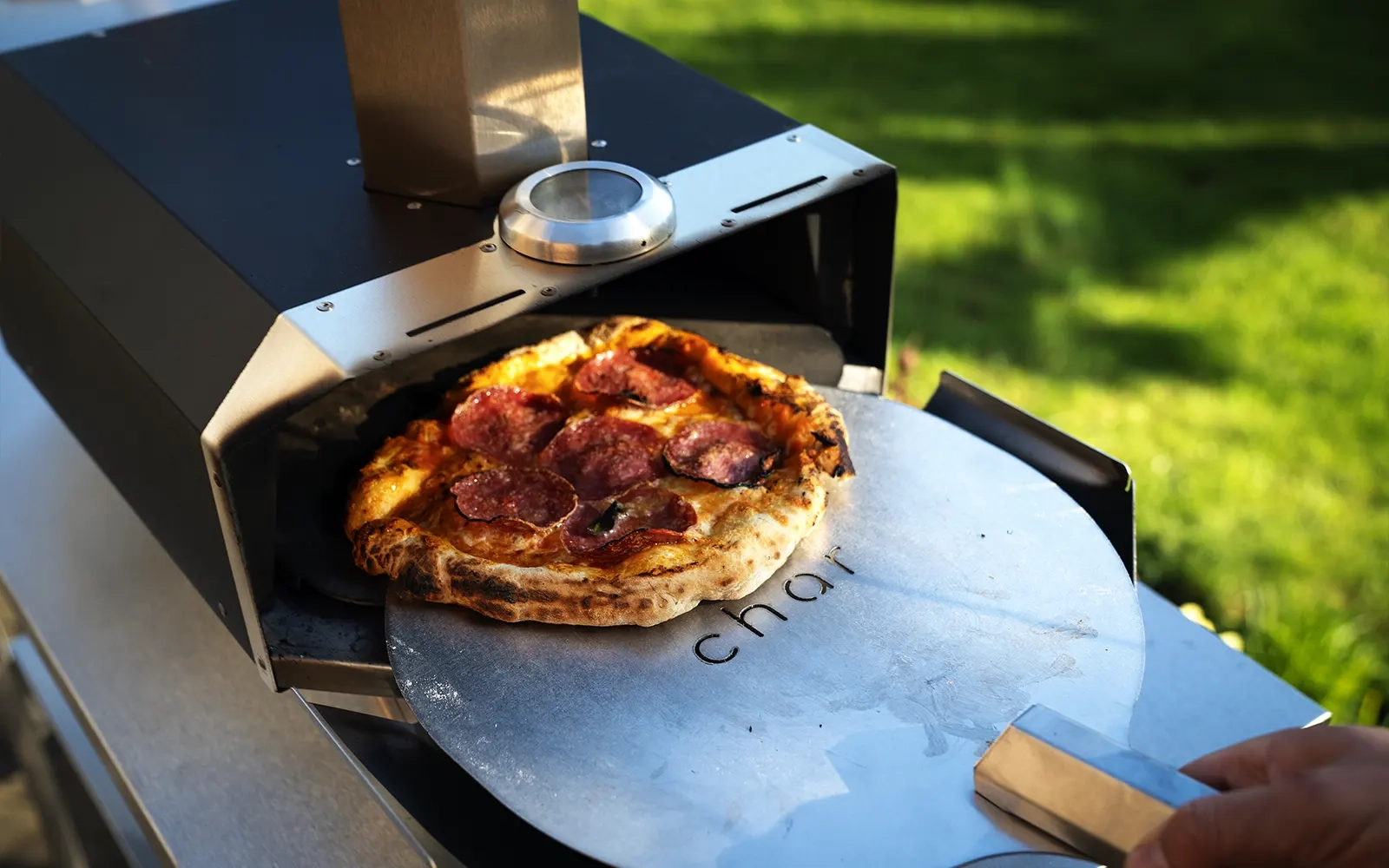 Close-up of a cooked pizza being taken out of a pizza oven on a pizza peel in a garden
